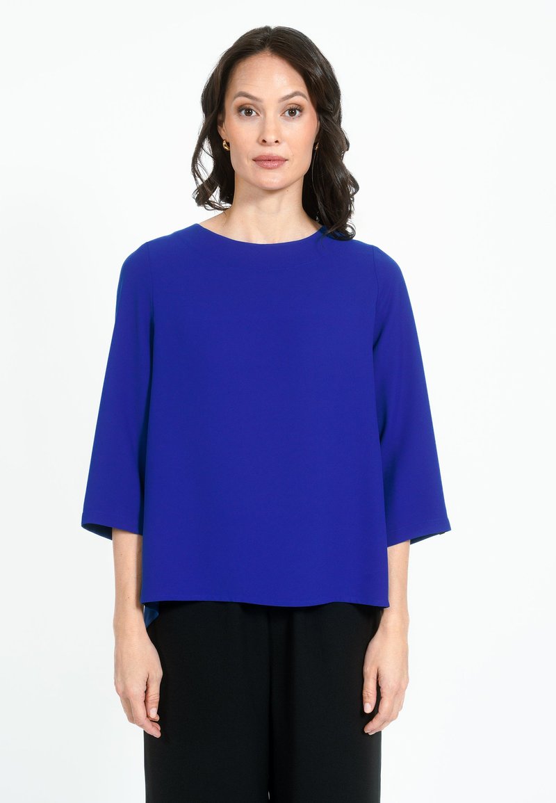 Blusa blu, morbida e ampia, con scollatura rotonda e maniche a tre quarti. Il tessuto appare liscio; abbinata a pantaloni neri.