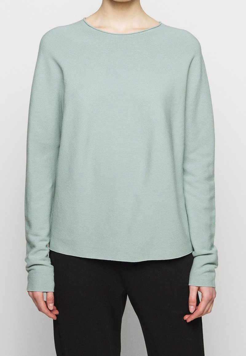 Personne portant un haut en maille vert clair à manches longues et un pantalon noir, debout devant un fond uni clair.
