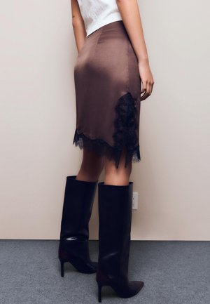 Rjava satenasta krilo z črnim čipkastim trakom na robu, združena z visokimi črnimi stiletto škornji, ki poudarjajo elegantno silhueto.