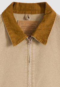 Ανοιχτό καφέ μπουφάν με γιακά από κοτλέ, κλείσιμο με φερμουάρ και υφασμένη ετικέτα με στοιχεία της μάρκας. Το ύφασμα φαίνεται ανθεκτικό και με υφή.