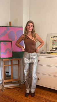 Καφέ τοπ με έναν ώμο, ασημί ψηλόμεσες υφασμάτινες παντελόνες, και μαύρες διαφανείς παπούτσια. Το φόντο περιλαμβάνει πολύχρωμο αφηρημένο πίνακα και έπιπλα.