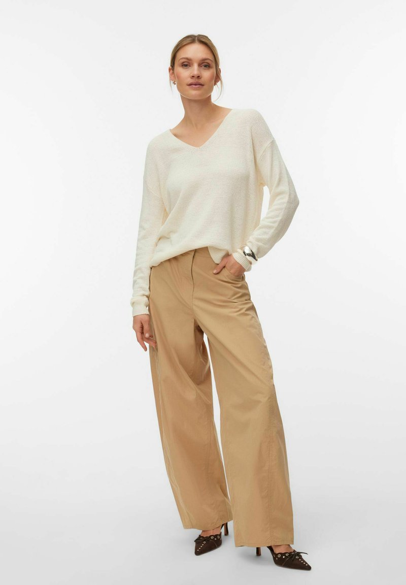 Femme portant un pull ample à col en V crème, un pantalon beige à jambes larges, et des talons pointus foncés, debout avec une main dans la poche.