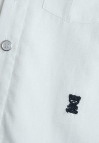 Chemise blanche boutonnée avec une poche, ornée d'une petite broderie d'ours noir sur la poitrine. Tissu texturé avec un design classique.