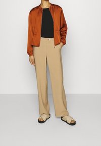Brændt orange satin bomberjakke, sort tætsiddende top, beige vide bukser og beige slidesandaler med sorte såler. Elastiske manchetter på jakken.