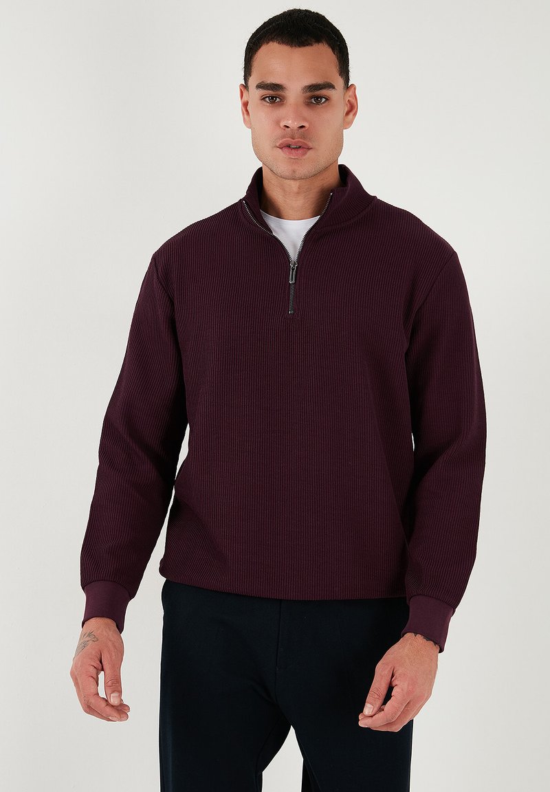 Suéter burdeos de canalé con cuello simulado con cremallera. Mangas largas y un corte relajado, combinado con una camiseta blanca debajo.