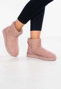 Persona que lleva botines de ante rosa claro con forro suave, combinados con jeans ajustados oscuros, de pie sobre un suelo blanco.