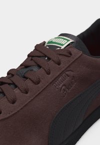 Κοντινό πλάνο καφέ σουέτ αθλητικού παπουτσιού Puma με κορδόνια στο ίδιο χρώμα, μαύρη ρίγα από δέρμα και πράσινο λογότυπο Puma στη γλώσσα.