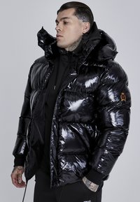 Chaqueta acolchada negra con material brillante, capucha de gran tamaño y múltiples bolsillos. Presenta un parche de la marca en la manga izquierda.