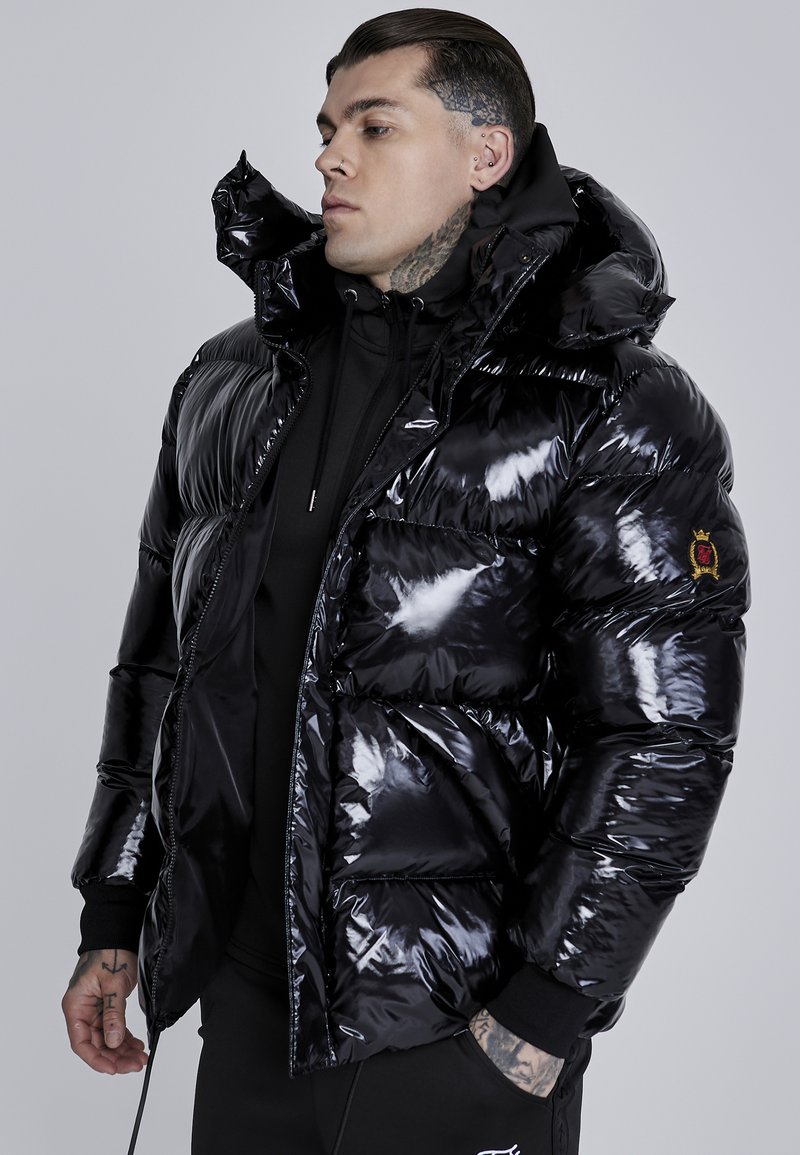 Chaqueta acolchada negra con material brillante, capucha de gran tamaño y múltiples bolsillos. Presenta un parche de la marca en la manga izquierda.