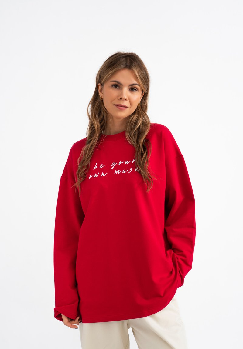 Czerwona, oversize bluza z miękkiego materiału, ozdobiona białym napisem "bądź swoją muzą". Długie rękawy i luźny krój, z okrągłym dekoltem.