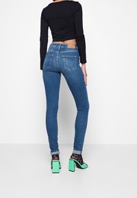 Jeans ajustados azules con bolsillos traseros, combinados con una camiseta negra corta y acentuados por sandalias de tacón verdes y calcetines negros con patrón.