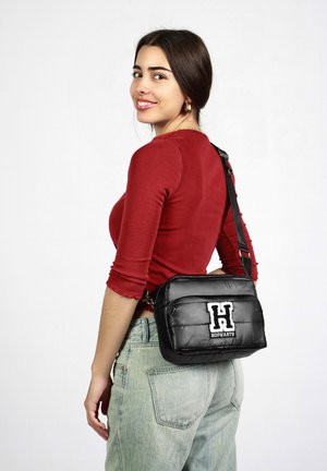 Černá, nadýchaná crossbody taška s nastavitelným popruhem, na přední straně s bílou vyšitou „H“ a textem „HOGWARTS“. 