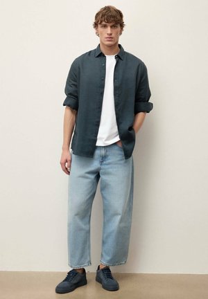 Jeune homme portant une chemise foncée retroussée sur un t-shirt blanc, un jean à jambes larges bleu clair et des baskets foncées, debout contre un mur uni.