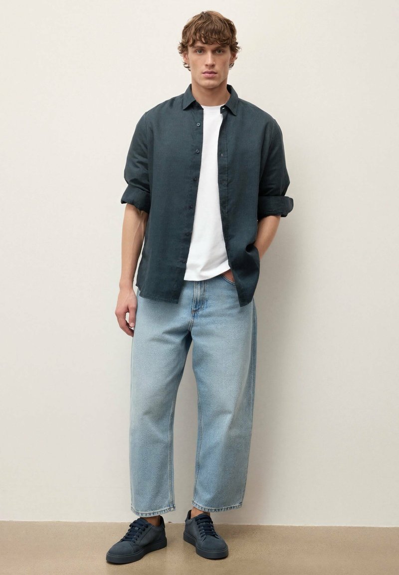 Jeune homme portant une chemise foncée retroussée sur un t-shirt blanc, un jean à jambes larges bleu clair et des baskets foncées, debout contre un mur uni.