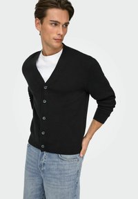 Cardigan nero realizzato in un tessuto a maglia fine con scollo a V, caratterizzato da sei bottoni rotondi sul davanti e maniche lunghe. Abbinato a jeans in denim chiaro.