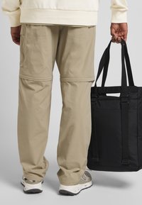 Beige lichtgewicht broek met zichtbare naden en cargozakken, gecombineerd met een zwarte draagtas. De schoenen zijn licht en hebben een textuurontwerp.