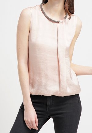 Woman wearing a sleeveless light pink blouse with a decorative collar and black pants, with one hand resting on her hip.