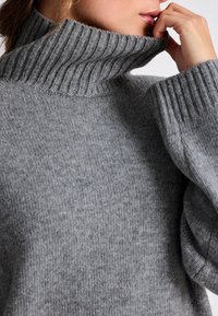 Jersey de cuello alto gris con textura acanalada y ajuste holgado. Textura suave, mangas anchas y un cuello alto que crean un aspecto acogedor.