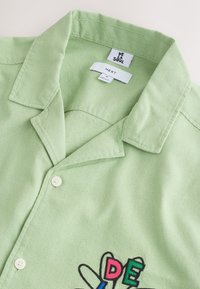 Ljusgrön skjorta med knappar och krage, medelstorleksetikett och färgglad "DE LA SOUL" broderad text nära den nedre kanten.