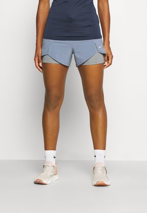 Shorts de sport courts en bleu et gris, avec un design superposé, des poches latérales et un logo Nike. Portés avec des chaussettes blanches et des baskets claires.