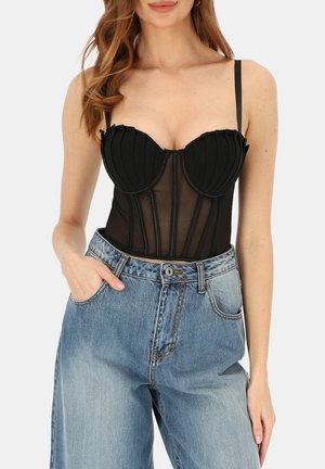 Kobieta w czarnym koronkowym gorsie i wysokich dżinsach z podwyższonym stanem, z jedną ręką w kieszeni, na jednolitym tle.