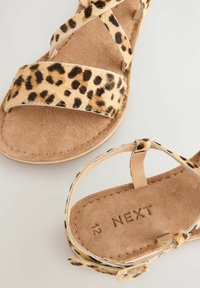 Sandalen mit Leopardenmuster, gekreuzten Riemen, einer weichen Beigefarbenen Wildleder-Innensohle und einem braunen Absatz. Mit einem goldenen metallischen Schnallendetail.