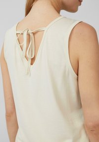 Cremefarbene ärmellose Bluse mit V-förmigem Rücken und einer Bindeschließe. Weicher Stoff mit sichtbaren Nahtakzenten.