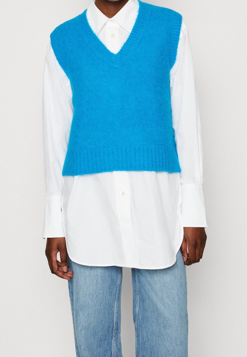 Personne portant un gilet sans manches en tricot bleu vif par-dessus une longue chemise blanche boutonnée et un jean bleu clair.