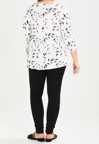 Top bianco con motivo di uccelli neri, maniche 3/4, abbinato a pantaloni neri aderenti e scarpe basse con dettagli decorativi.