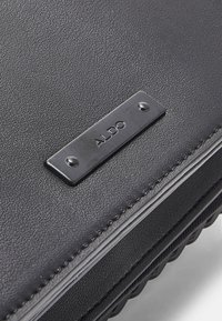 Svart syntetiskt läderväska med en texturerad yta, med en metallisk rektangulär ALDO-logoplåt och kontrasterande sömmar.