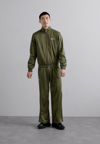 Ελαιόχρουν αθλητική φόρμα με ζακέτα με φερμουάρ και ελαστική μέση. Κατασκευασμένη από λείο ύφασμα. Μαύρα υποδήματα συμπληρώνουν την αθλητική εμφάνιση.