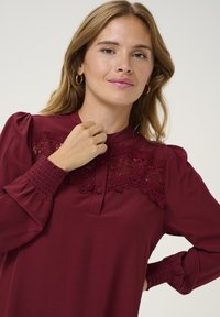 Burgundska bluzica z visokim čipkastim ovratnikom in smockanimi manšetami. Ima henley dekolte z gumbi in dolge napihnjene rokave.