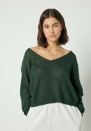 Un pull doux vert forêt avec un décolleté en V profond et une coupe ample. La texture semble légèrement transparente, associé à un pantalon gris clair.