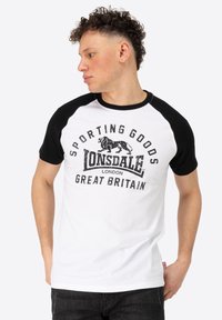 Balts un melns raglānu T-krekls ar grafikā attēloto uzrakstu "Lonsdale London" un "Sporting Goods" detaļām. Īsas piedurknes, kokvilnas audums.