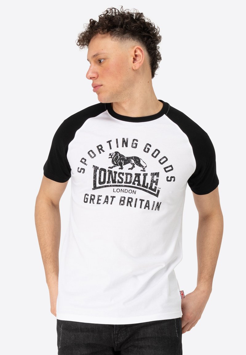 Balts un melns raglānu T-krekls ar grafikā attēloto uzrakstu "Lonsdale London" un "Sporting Goods" detaļām. Īsas piedurknes, kokvilnas audums.