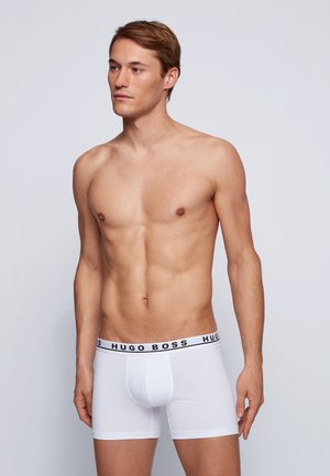 Weiße Baumwoll-Boxershorts mit einem schwarzen "HUGO BOSS"-Bund, taillierte Passform und glatter Textur. Das Model posiert mit neutralem Ausdruck.