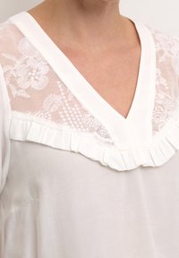 Blusa blanca con escote en V, que presenta un panel de encaje transparente con patrones florales y un borde rizado a lo largo del escote. Textura suave.