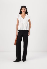 White V-neck blouse with ruffle sleeves, paired with black wide-leg trousers. The model holds a brown handbag and wears pointed black shoes.