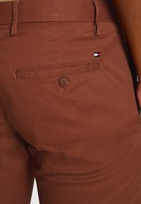 Brown trousers featuring a textured fabric, a buttoned back pocket, and a logo patch with red, white, and blue colors on the waistband.