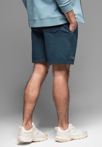 Pantalones cortos de color navy hechos de una tela suave, con bolsillos laterales y una etiqueta de logo. Combinados con zapatillas de deporte de color claro.