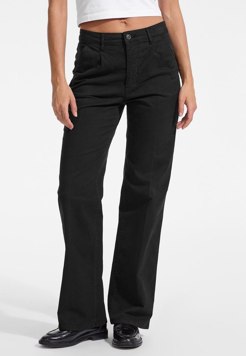 Crne hlače ravnog kroja izrađene od pamuka s kopčanjem na gumb, imaju bočne džepove i glatku teksturu. Nosene s tamnim cipelama.