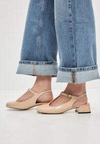 Beige patent leather T-strap kengät, joissa on matala lohko korko ja leikattu sivut. Yhdistettynä vaalean sinisiin farkkuihin, joissa on käännetyt lahkeet.