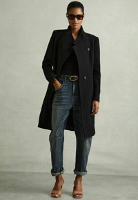 Abrigo negro largo con un diseño de doble botonadura, combinado con jeans azul enrollados y tacones nude. Los accesorios incluyen gafas de sol oscuras y un cinturón.