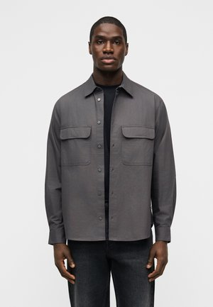 Άνδρας που φοράει γκρι πουκάμισο με κουμπιά και δύο τσέπες στο στήθος πάνω από μαύρο πουκάμισο και σκούρο τζιν, στέκεται μπροστά σε απλό φόντο.