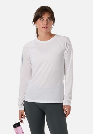 Kvinde med brunt hår iført en hvid langærmet trøje og mørke leggings holder en lyserød vandflaske mod en hvid baggrund.