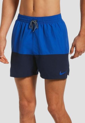 Uomo che indossa pantaloncini da nuoto Nike bicolore blu e navy con vita con coulisse, mostrato dalla metà del busto alla metà della coscia su sfondo neutro.