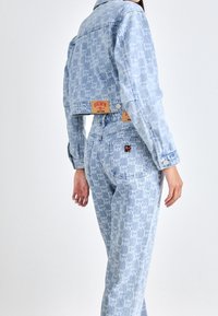 Ljust blå denimjacka och matchande jeans med ett upprepande logomönster. Jackan är kort, med märkta lappar på midjan.