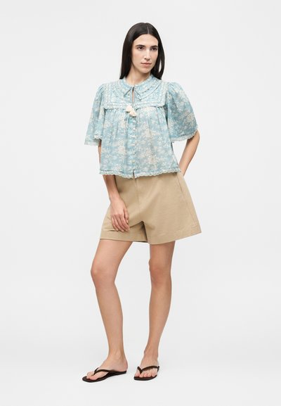 Γυναίκα που φοράει ανοιχτό μπλε φλοράλ μπλούζα με φουσκωτά μανίκια και μπεζ σορτς, στέκεται με το ένα χέρι στην τσέπη και μαύρες σαγιονάρες.