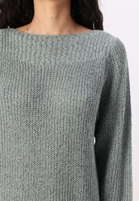 Femme portant un pull tricoté gris clair avec un motif côtelé texturé et des manches longues sur un fond clair uni.