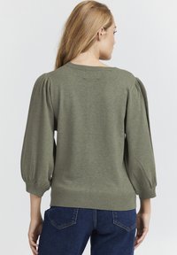 Maglione verde lavorato a maglia con maniche a sbuffo, polsini e orlo a costine, presenta una vestibilità comoda e una piacevole texture. Indossato con jeans blu.